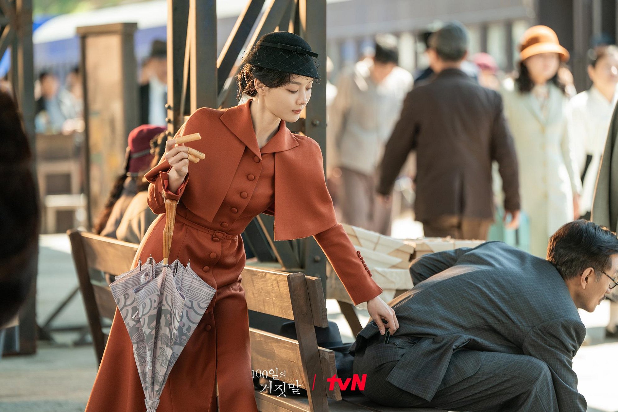 Lee Ga-kyung in a rust-colored period coat and hat at a crowded station in 100 Days of Deception